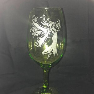 May include: A green glass wine glass with an etched design of a fairy with wings. The fairy is holding a flame in her hand.