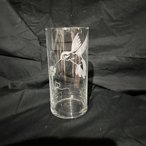 May include: A clear glass vase with an etched design of a hummingbird in flight. The vase is on a black background.