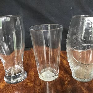 May include: Three clear glass drinking glasses. The glass on the left has a wide base and a tall, slender body. The glass in the center has a straight, tall body and a wide base. The glass on the right has a rounded body and a textured surface.