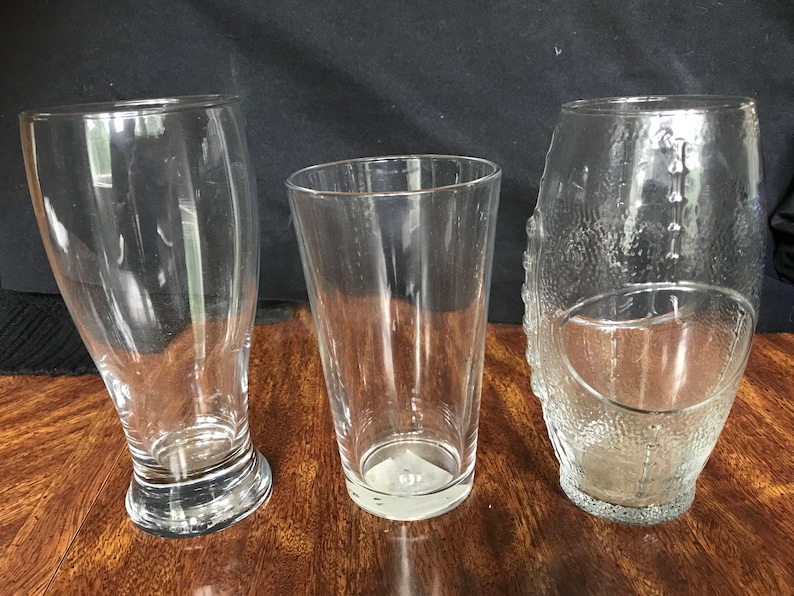 May include: Three clear glass drinking glasses. The glass on the left has a wide base and a tall, slender body. The glass in the center has a straight, tall body and a wide base. The glass on the right has a rounded body and a textured surface.