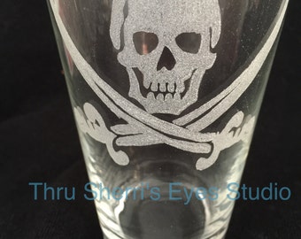 May include: A clear glass pint glass with an etched design of a skull and crossbones. The design is in a frosted white color.