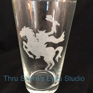 May include: A clear glass pint glass with an etched design of a horse and rider. The text "Thru Sherri's Eyes Studio" is visible at the bottom of the glass.