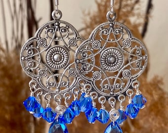 Op de afbeelding: Zilverkleurige filigraan oorbellen met een bloemmotief. Elke oorbel heeft een centraal medaillon en is versierd met bungelende blauwe kristallen kralen. De oorbellen hangen aan zilveren haken, waardoor een delicaat en opvallend accessoire ontstaat.
