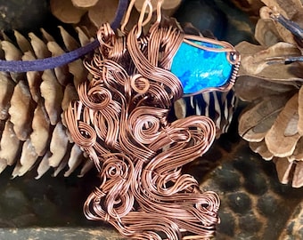 Op de afbeelding: Een met draad omwikkelde hanger met een felblauwe steen gezet in ingewikkelde, wervelende koperdraadspiralen. De hanger hangt aan een donkerpaars koord en toont een gedetailleerd metaalontwerp dat de levendige blauwe edelsteen omlijst.