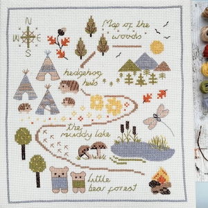 Puede incluir: Un patrón de punto de cruz de un mapa del bosque con una brújula, árboles, un lago, una fogata y animales como erizos y osos. El texto en el mapa dice "Map of the woods", "hedgehog huts", "the muddy lake", y "little bear forest".