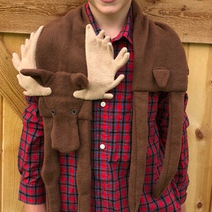 May include: A brown fleece moose scarf with a red and black plaid shirt underneath. The scarf has a moose head with large antlers and two long legs.