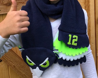 Puede incluir: Una bufanda de forro polar azul con un diseño de búho verde y blanco y el número "12" bordado en el extremo. La bufanda la lleva puesta un joven que hace un pulgar hacia arriba.
