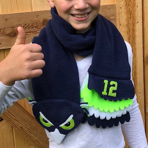 May include: A blue fleece scarf with a green and white owl design and the number "12" embroidered on the end. The scarf is worn by a young person with a thumbs up.