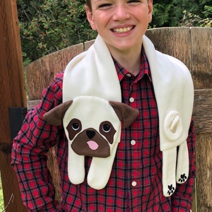 May include: A white fleece scarf with a brown and white pug face design. The scarf is worn by a young person wearing a red and black plaid shirt.