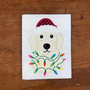 May include: White dog wearing a red and black plaid Santa hat with a white trim and a string of colorful Christmas lights around its neck.