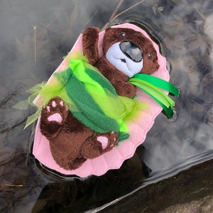 Op de afbeelding: Een bruine pluche otter die een groen en roze tutu draagt met groene lintjes, drijvend in een plas water.