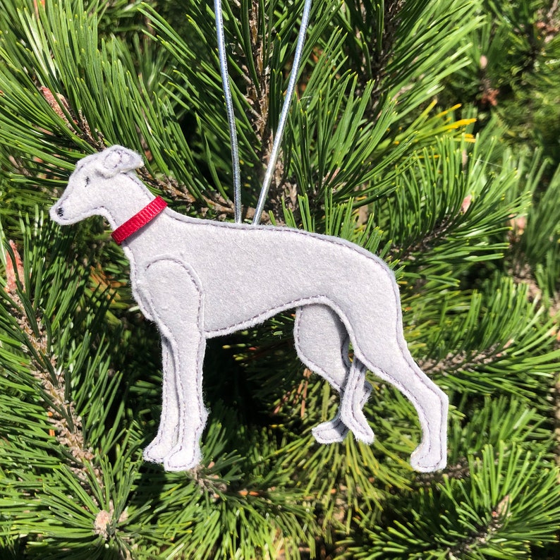 May include: A gray felt dog ornament with a red collar, hanging from a silver string. The dog is a greyhound, with detailed stitching. The ornament is displayed against a backdrop of green pine needles, suggesting a holiday theme.