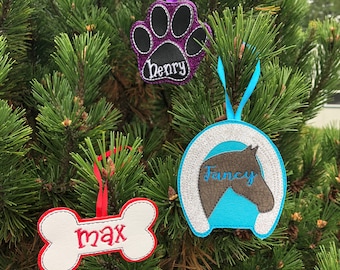May include: Three Christmas ornaments with personalized names. A purple paw print ornament with the name "Henry", a blue horseshoe ornament with the name "Fancy", and a white bone ornament with the name "Max".