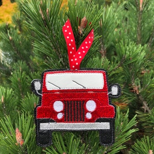 May include: Red glitter Jeep bauble with a white windscreen and white headlights. The bauble is hanging from a red and white polka dot ribbon.