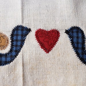 Puede incluir: Dos pájaros de cuadros azules y blancos con detalles marrones se miran con un corazón rojo en el medio. Los pájaros y el corazón están cosidos sobre una tela beige.