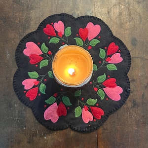 Peut inclure: Un dessous de plat en feutre noir avec un bord festonné et un motif floral composé de cœurs roses et rouges. Une bougie allumée est placée au centre du dessous de plat.