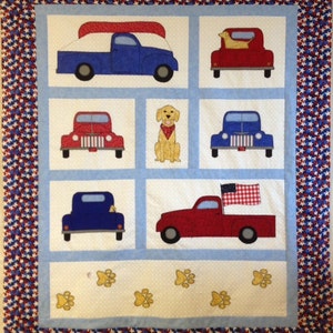May include: A quilt with a blue and white background featuring red, blue, and white trucks and a golden retriever dog. The quilt has a red, white, and blue star border. The quilt also features paw prints.