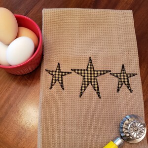 May include: A brown waffle weave kitchen towel with three black and yellow gingham stars sewn on. The towel is lying on a wooden surface.