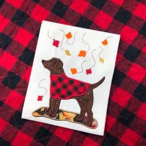 May include: Embroidered design of a brown dog wearing a red and black plaid jacket with falling leaves around it. The design is on a white fabric background.