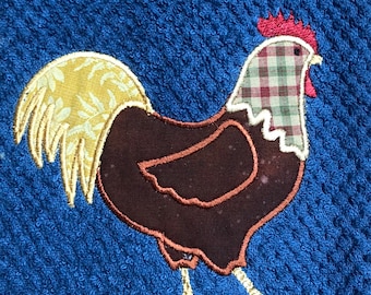 May include: A blue kitchen towel with a decorative rooster applique. The rooster features a brown body, a plaid head, and a yellow and white patterned tail. The rooster's comb is red, and its feet are yellow.