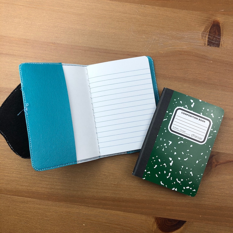 May include: An open, lined notebook with a turquoise cover sits next to a small composition book with a green speckled cover. The turquoise notebook is open to a blank page. The composition book has a white label.