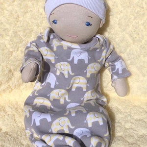 May include: A handmade cloth doll wearing a gray and yellow elephant print sleeper and a white and gray knit hat. The doll has blue eyes and a stitched smile.