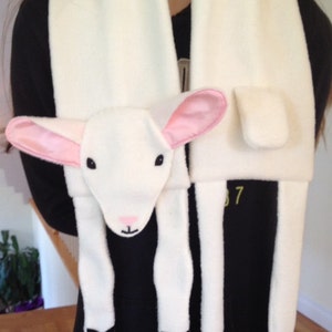 May include: White fleece scarf with a white lamb face and pink ears. The lamb has black hooves and a pink nose. The scarf is worn around a person's neck.