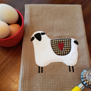 May include: A brown waffle weave kitchen towel with a white sheep applique. The sheep has black eyes and a black nose. The sheep has a red heart on its back with a black and white checkered background.