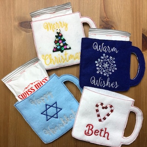 Könnte beinhalten: Vier Filz-Becherwärmer mit gestickten Designs und Text. Die Becherwärmer sind in Blau, Weiß und Gelb. Der Text auf den Becherwärmern lautet "Merry Christmas", "Warm Wishes", "Happy Hanukkah" und "Beth".
