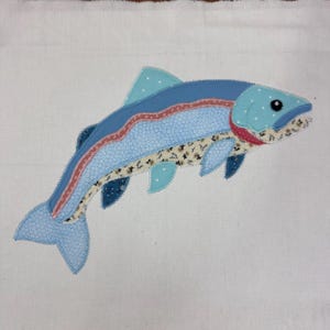 May include: A fabric appliqué of a fish featuring a light blue body with white polka dots, a dark blue top section, a floral patterned midsection, and a pink stripe. The fish has a dark blue eye and dark blue fins on a plain white background.