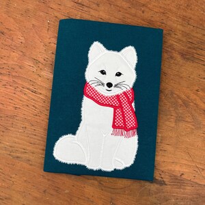 May include: A white arctic fox wearing a red and white checkered scarf, embroidered on a dark teal fabric.