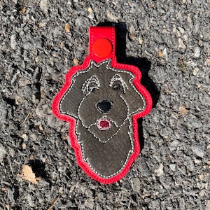 May include: A red leather keychain with a gray embroidered dog head. The dog has a black nose and a pink tongue.