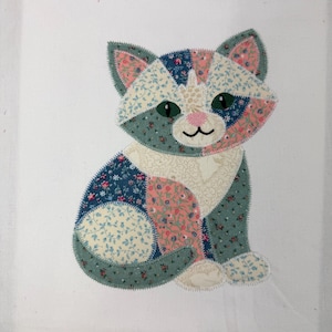 May include: A cat-shaped fabric appliqué featuring a patchwork design of various floral and geometric patterns in shades of blue, pink, green, and cream. The cat has dark green eyes, a pink nose, and a black stitched mouth on a white background.