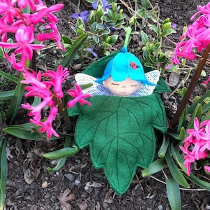 May include: A handmade felt fairy doll sleeping in a green leaf bed. The fairy has a blue hat with a ladybug on top and white wings. The fairy is surrounded by pink hyacinth flowers.