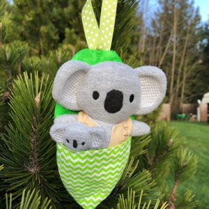 May include: A gray koala plush toy with a smaller gray koala plush toy in its pouch. The larger koala is wearing a tan vest with brown stitching. The plush toys are hanging from a green and white polka dot ribbon on a green tree branch.