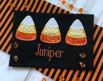 May include: Black fabric with three embroidered candy corn designs and the name "Juniper" in white thread.