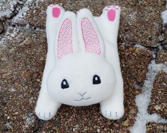 May include: A white plush bunny with pink ears and black eyes. The bunny is lying on its back with its paws in the air.