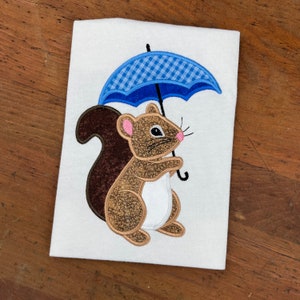 May include: Embroidered design of a brown squirrel holding a blue and white checkered umbrella on a white t-shirt.