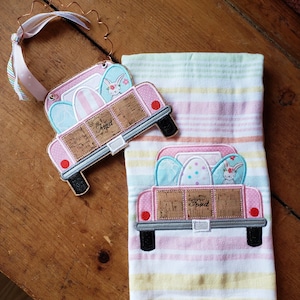 May include: Two pink pickup trucks with a wood bed are filled with Easter eggs and a bunny. The trucks are embroidered on a striped kitchen towel. The text "Happy Easter" is on the back of the trucks.