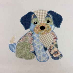 May include: A patchwork dog applique in a sitting position. The dog's ears are denim blue, and its body is composed of floral, gingham, and patterned fabrics in shades of blue, pink, beige, and green. The dog has black eyes and a black nose.