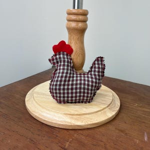May include: A red and black checkered fabric chicken figurine with a red comb and wattle. The chicken is sitting on a wooden base.
