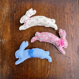 May include: Three handmade fabric bunny rabbits. One is white with pink floral print, one is pink with white floral print, and one is blue with white gingham print. Each bunny has a pink bow.