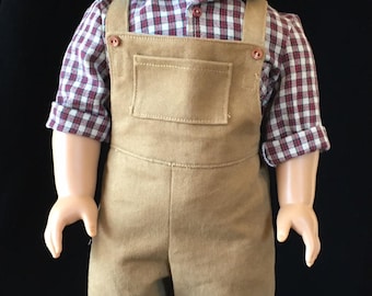 Puede incluir: Una muñeca con una camisa a cuadros rojos y blancos y un mono marrón. La muñeca tiene el pelo castaño y los ojos verdes.
