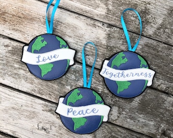 Op de afbeelding: Drie blauwe vilten ornamenten in de vorm van de aarde met witte banners. De banners hebben de woorden "Love", "Togetherness" en "Peace" geborduurd.