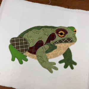May include: A fabric applique of a frog centered on a white background. The frog is constructed from various patterned textiles including green floral prints, brown and red plaid, and solid green fabric, all joined with visible blanket stitching.
