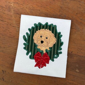 May include: Embroidered design of a dog's head peeking out of a green and white striped wreath with a red bow.