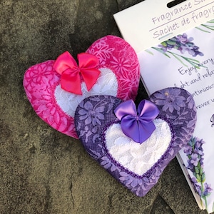 May include: Two heart-shaped fabric sachets with lace trim and satin bows. The pink sachet has a floral pattern and a pink bow. The purple sachet has a floral pattern and a purple bow. The sachets are on a brown surface.