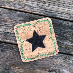 Może przedstawiać: Podkładka korkowa z czarną gwiazdą w środku i zielono-czerwoną haftowaną ramką. Podkładka leży na drewnianej powierzchni.
