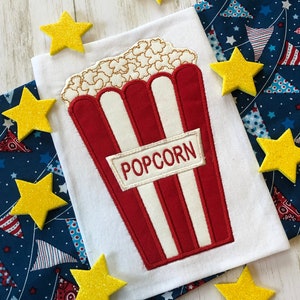 Puede incluir: Una camiseta blanca con un cubo de palomitas de maíz rojo y blanco bordado en la parte delantera. El cubo tiene la palabra "POPCORN" escrita en él. La camiseta está rodeada de estrellas de brillantina amarillas.