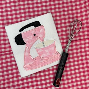 May include: A pink stand mixer with a white bowl and a black handle. The mixer is on a pink and white checkered background.